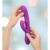 A person's hands holding a purple Femme Funn Ultra Rabbit vibrator by Femme Funn against a softly blurred background with pastel lights, showcasing its powerful vibrations and uniquely curved cylindrical shape with two protruding sections designed for exceptional dual G-spot stimulation.